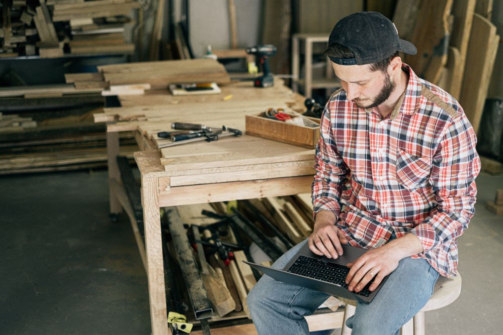 A carpenter on a stool in a workshop, laptop on his knee