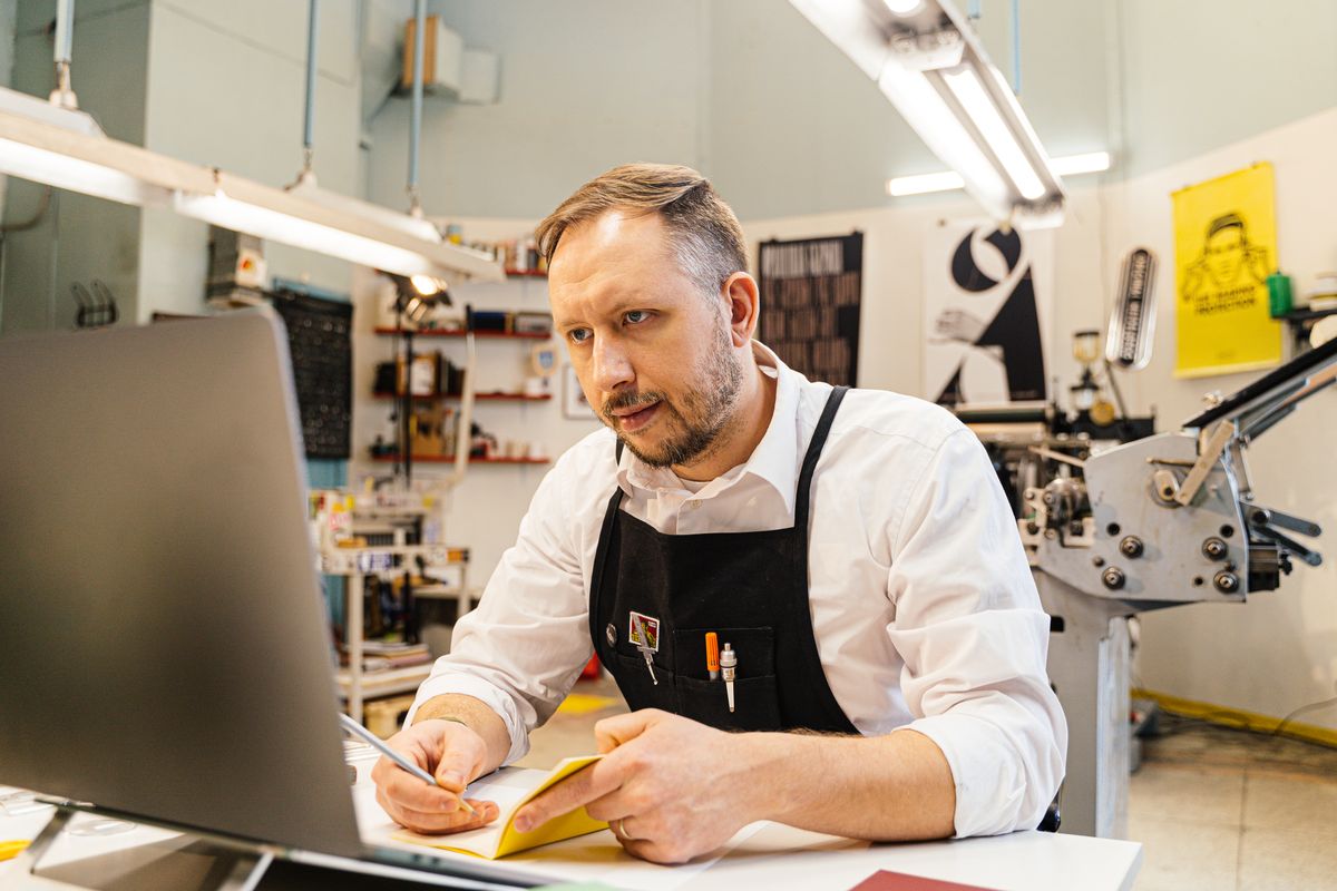 A print shop owner in an apron working at a laptop with a notebook